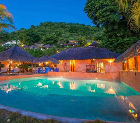 Luxurious resort pool at twilight with thatched roofs and hillside backdrop