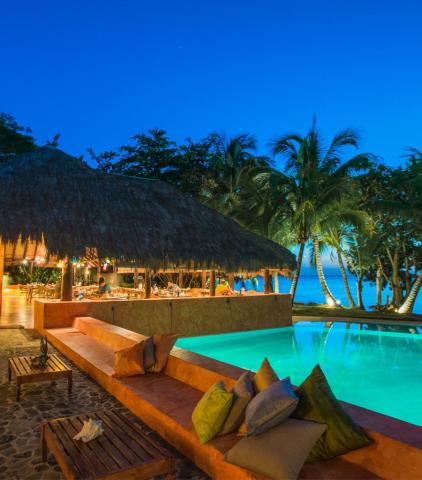 Tropical resort with pool, thatched bar, and ocean view at dusk