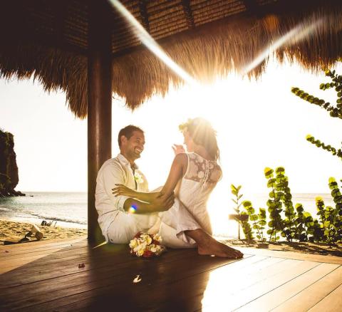 Joyful couple in white laughing under a thatched roof by the beach at sunset