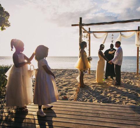 Beach wedding ceremony with bride and groom under arch, flower girls watching.