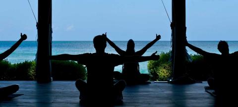 Three people meditating on a deck overlooking a serene ocean with lush greenery