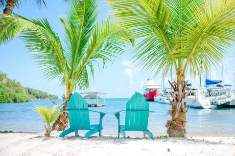 Turquoise Adirondack chairs on beach with boats and palm trees in background.