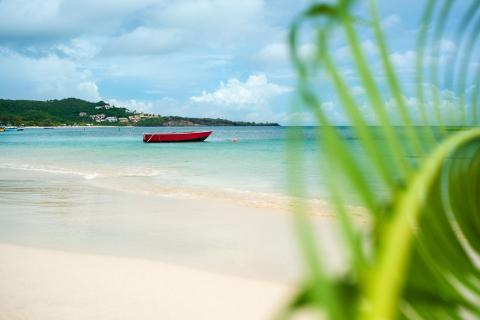 Tranquil beach scene with moored red boat, turquoise water, and lush hill backdrop.