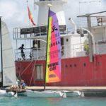 Colorful harbor scene with a red-hulled ship, sailboat, and worker on deck.