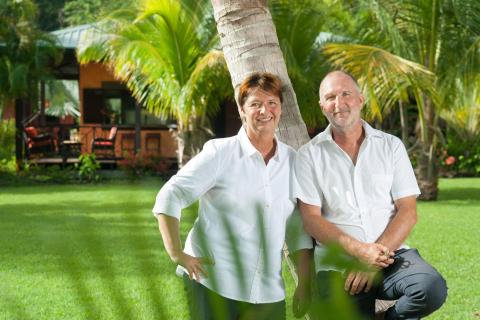 Middle-aged couple smiling in garden by tropical house, dressed in white.