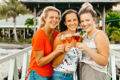 Three women toasting with pink drinks on a tropical boardwalk