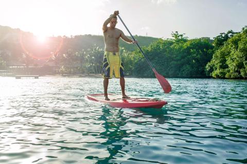 Man paddleboarding at sunset on calm lake, lush background.