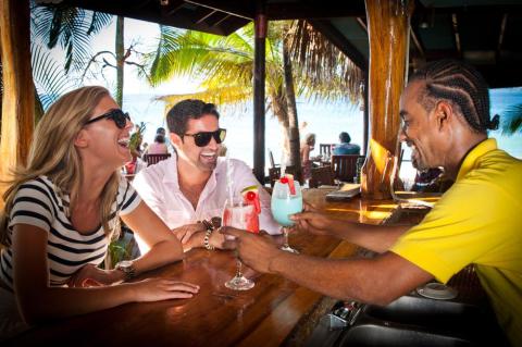 Friends laugh at beach bar with bartender serving drinks, sunny day.