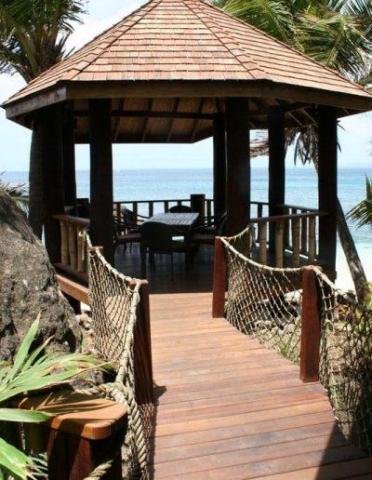 Wooden seaside gazebo with shingled roof, deck, and tropical plants, perfect for dining.