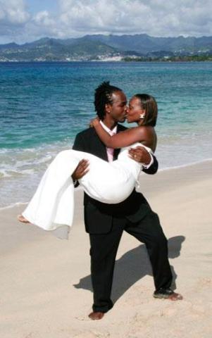 Joyous couple in wedding attire kissing on a sunny beach with mountain backdrop.