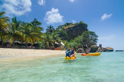 Two kayakers in tropical waters near palm-lined beach with cliff-top structure.