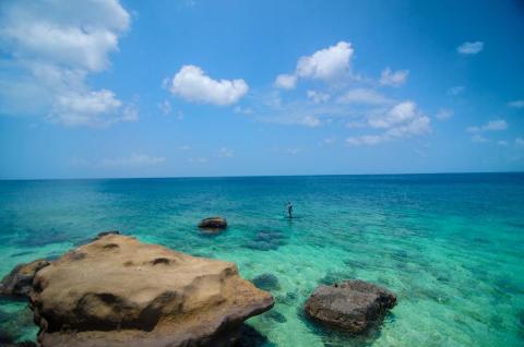 Paddleboarder in turquoise waters near rocky shore under clear sky.