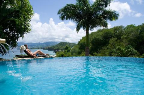 Person relaxing in infinity pool with hillside and ocean view, sunny day.