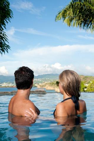 Couple in infinity pool overlooking mountains under blue skies, tropical setting.