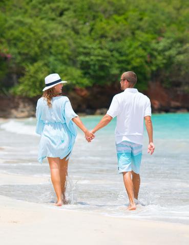 Couple walking on beach, man in white shirt, woman in blue dress and hat by ocean.
