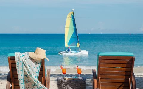 Tranquil beach setting with lounge chairs, tropical drinks, and a colorful sailboat.