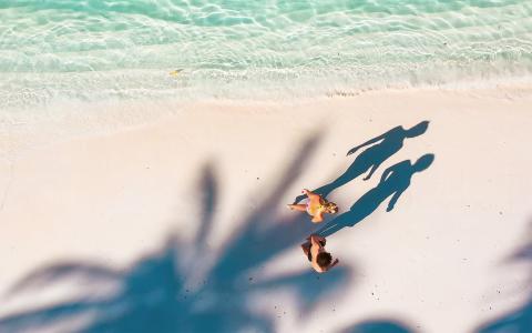 Aerial view of sandy beach with clear turquoise waters, two people, and palm shadow