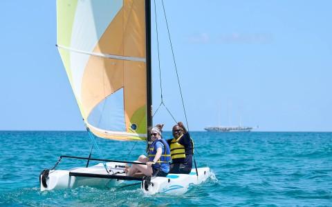 Two people sailing on a small catamaran with a yellow sail in clear blue sea