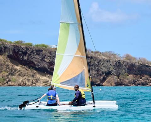 Two people and a dog sailing near a cliff on a sunny day