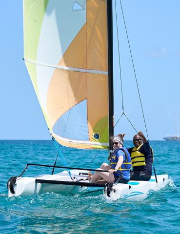 Two people enjoy sailing on a vibrant catamaran in clear blue waters under sunny skies.