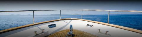 Yacht deck with polished rail, cleats, and hatch overlooking calm sea and distant hills