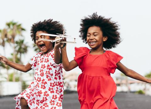 Two girls playing outdoors with airplane, in floral and red dresses, joyful.