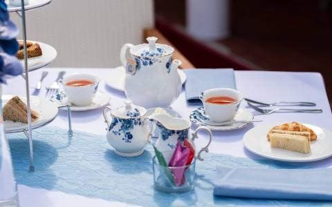 Outdoor tea party setup with porcelain tea set, cakes, and sandwiches on table.