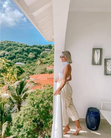 Woman in white top on balcony overlooking lush landscape on sunny day.