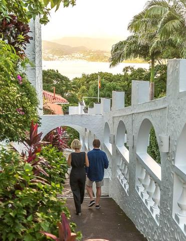 Couple walks under white archway with coastal town view in lush setting.