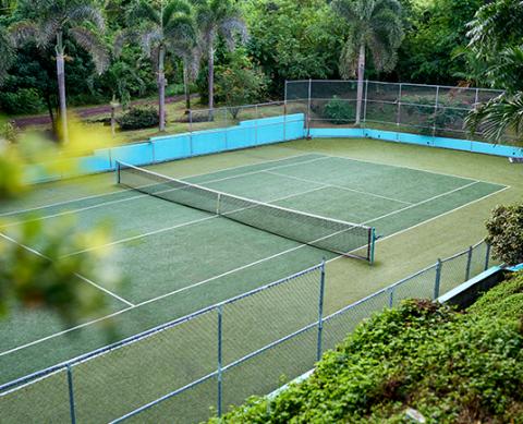Tropical outdoor tennis court with synthetic surface encircled by lush greenery and high fences.