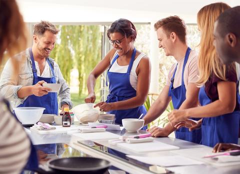 Adults in a cooking class wearing blue aprons, laughing as they bake in a sunlit kitchen.