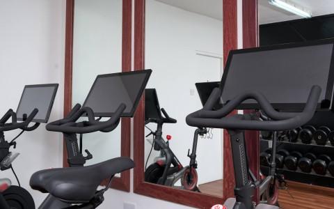 Indoor gym with stationary bikes and digital displays, mirrored wall.