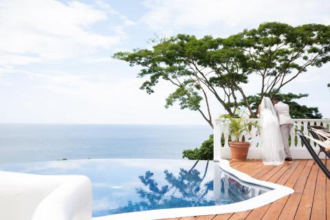 Newlyweds on balcony overlooking sea and infinity pool at tropical resort