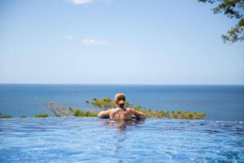 Person in infinity pool overlooking ocean, clear sky above, peaceful scene.
