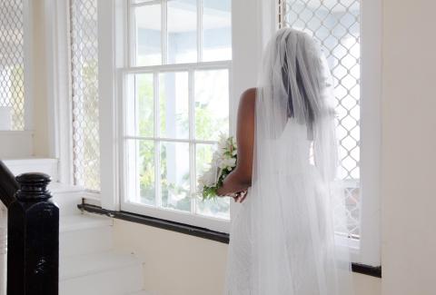 Bride in gown by window, bouquet in hand, traditional room setting.