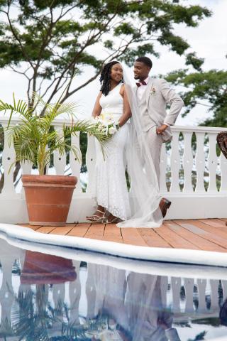 Newlyweds on deck in wedding attire, joyful pose by tranquil water backdrop.