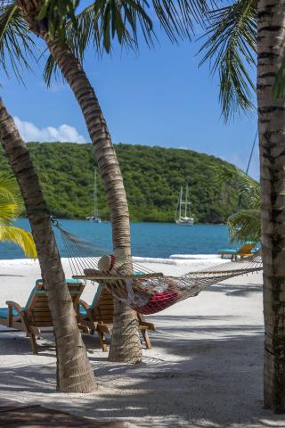 Relaxing beach scene with hammock, chairs, sailboats, and lush hills.