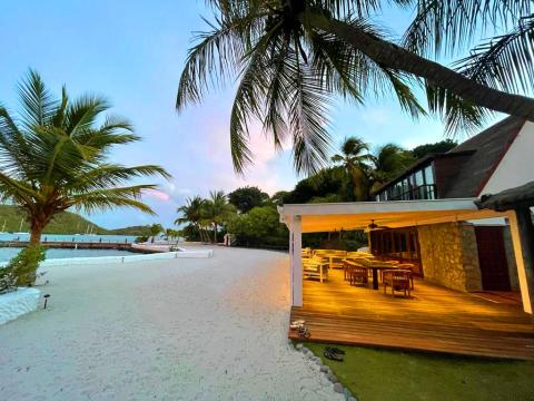 Twilight at beachside deck with tables, sandy shore, boats, and palm trees during twilight.