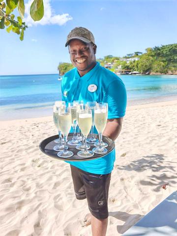 Man serving sparkling drinks on a beach, blue polo and cap, scenic backdrop