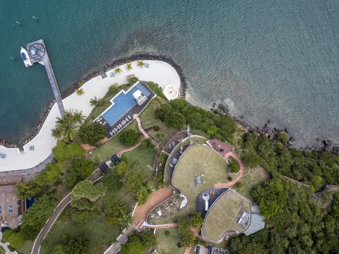 Aerial view of a resort with a sandy beach, pool, pier, and boat in a coastal setting.