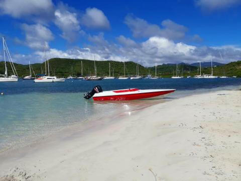 Red speedboat on sunny beach with sailboats, clear water, and green hills
