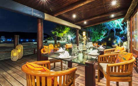 Outdoor dining setup on a wooden deck by the beach with illuminated trees and cozy lighting.