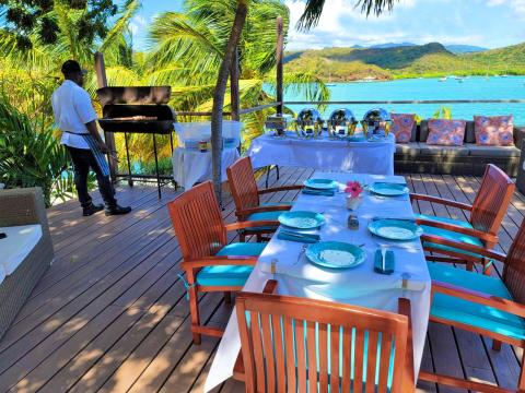 Outdoor dining setup with chef barbecuing on deck overlooking a scenic bay with sailboats.