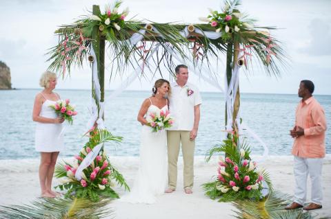 Beach wedding ceremony with couple, officiant, and bridesmaid under floral arch.