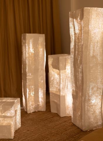 The image shows a set of illuminated, translucent sculptural objects of varying heights and shapes arranged on a carpeted floor