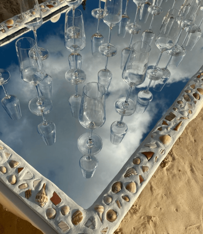 The image shows a creative display of wine glasses suspended above a reflective surface, which mirrors the blue sky and clouds, creating a dreamlike effect