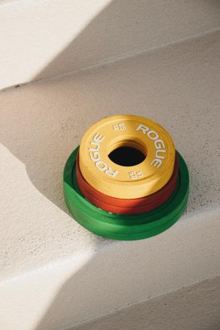 The image shows a stack of weightlifting plates laid on a textured floor, exposed to sunlight that casts a soft shadow