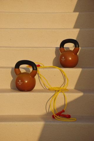 The image shows two brown kettlebells and a yellow jump rope placed on steps that are bathed in sunlight, creating a pattern of light and shadows across them