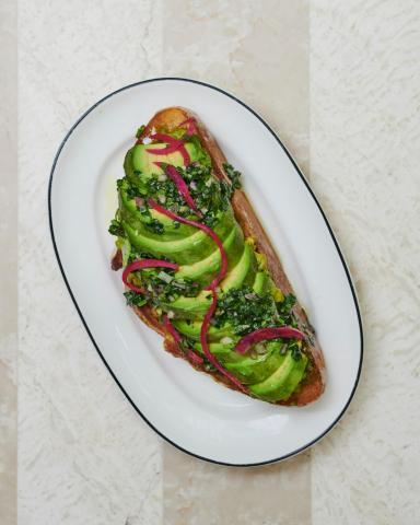 The image features a piece of toast topped artistically with avocado slices, a drizzle of pesto sauce, and possibly some red onion strands, all garnished with leafy chopped herbs that might be parsley or cilantro