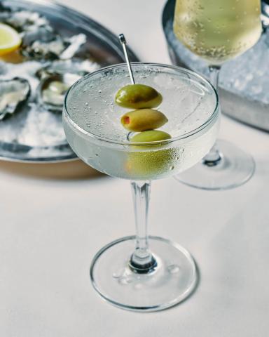 The image features a cocktail in a classic coupe glass, garnished with three green olives on a skewer